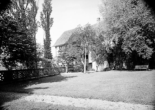 View over a lawn of buildings almost completely behind trees and bushes. On the left are black poplar trees. There is a wooden bench under one of the trees. Photo in black and white. The lens didn’t quite cover the picture circle.